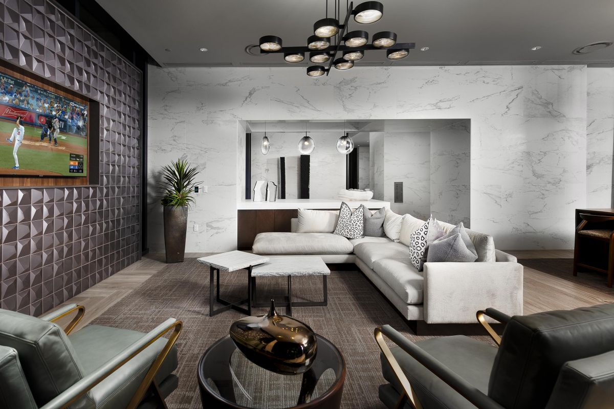 Modern living room with a marble accent wall, beige sectional sofa, geometric tables, and a wall-mounted TV displaying sports.