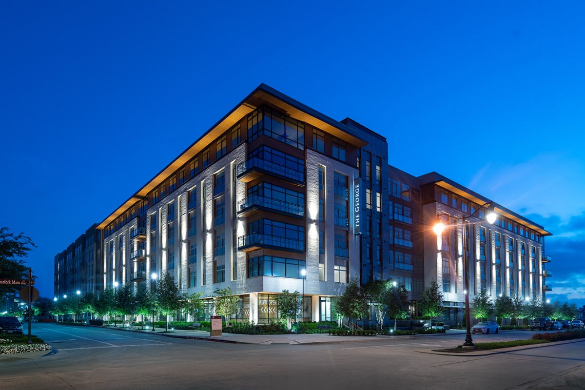 Modern apartment building with large windows and lights on at dusk, cars parked along the street.