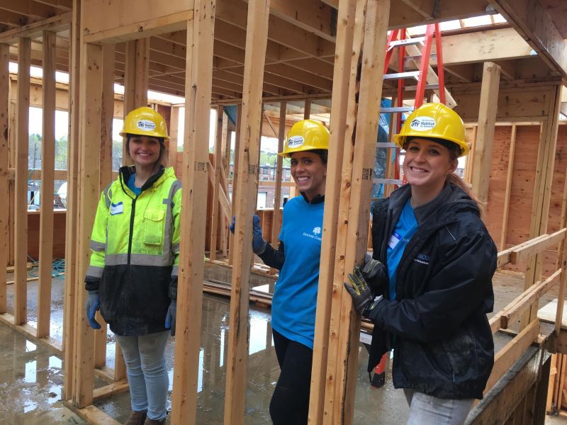 Three women in hard hats and gloves smile while working at a wooden house construction site.