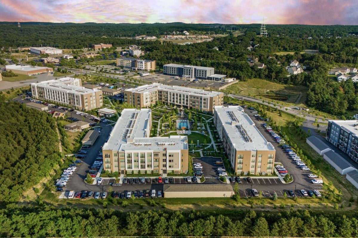 Aerial view of a large residential complex with parking, surrounded by trees and suburban buildings.