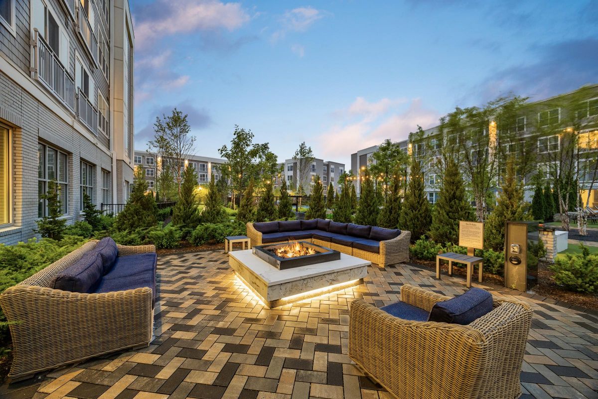 Outdoor patio with wicker seating surrounding a lit fire pit, framed by greenery and modern buildings.