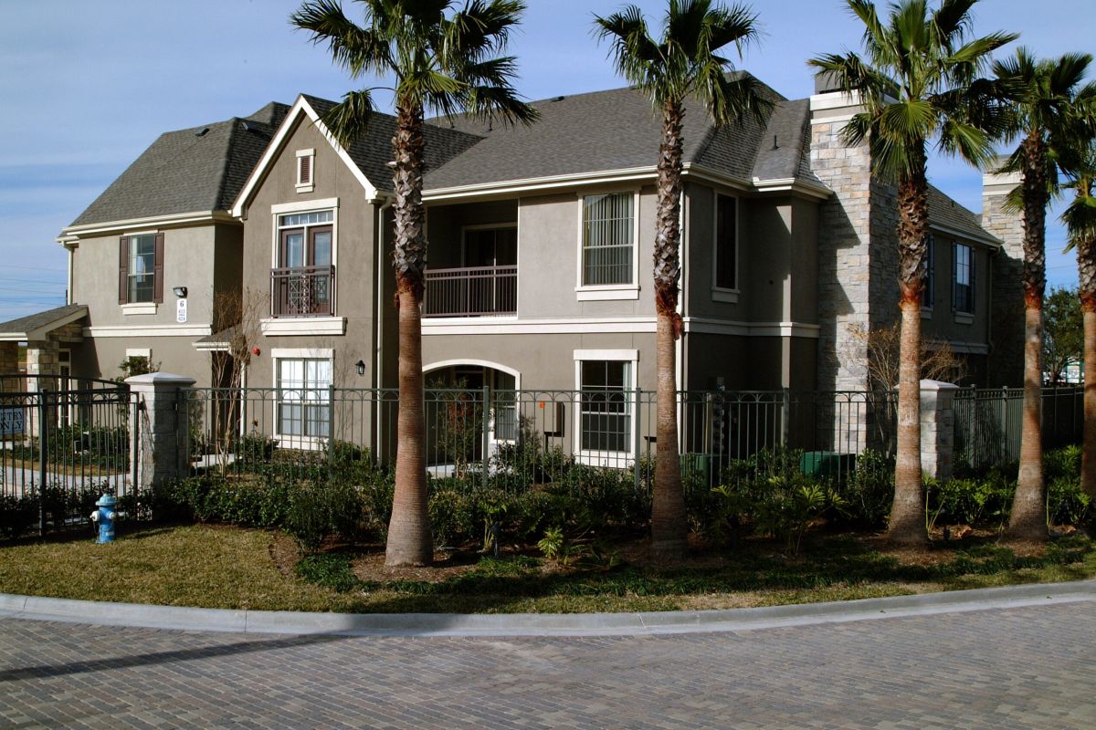 Two-story suburban house with gray exterior, palm trees, and a fenced yard seen from the street.