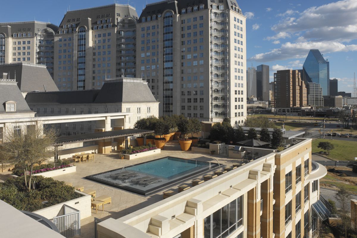 A rooftop pool area with seating, surrounded by tall buildings in a cityscape under a blue sky.