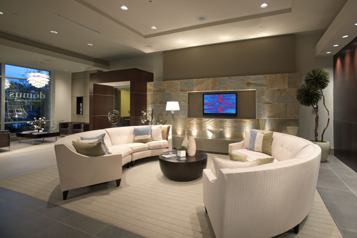 Modern hotel lobby with curved beige sofas, a round coffee table, indoor plants, and a wall-mounted TV.