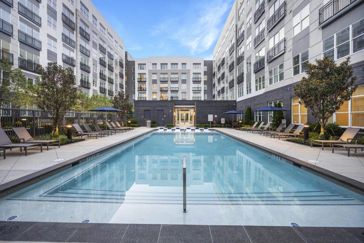 Modern apartment complex with a central pool, lounge chairs, and surrounding greenery under a clear blue sky.