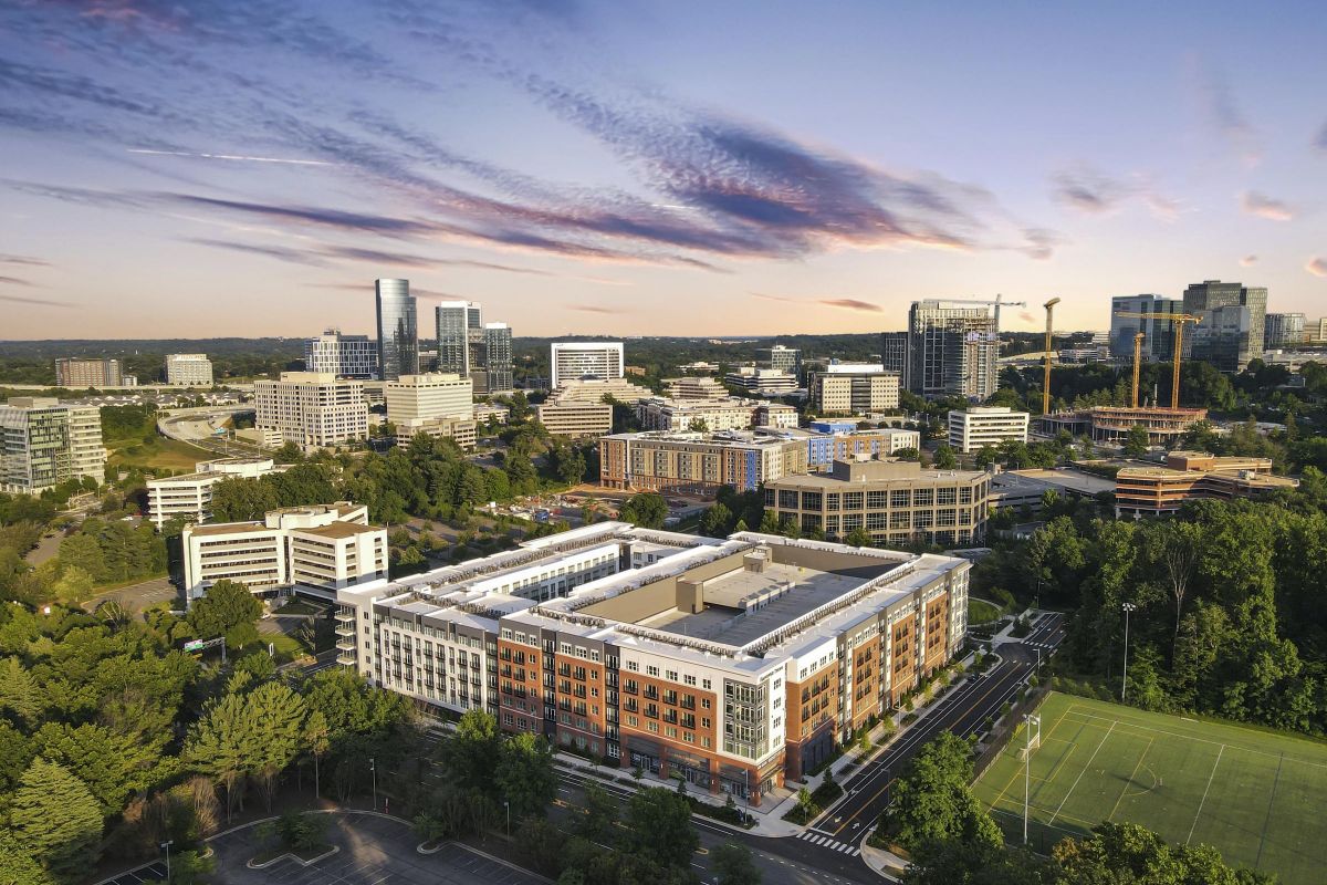 Aerial view of a cityscape with modern buildings, green spaces, and dramatic sky at sunset.