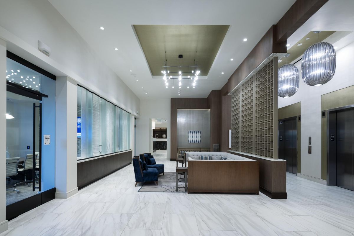 Modern hotel lobby with elevators, blue chairs, a desk, and decorative lighting on a white marble floor.