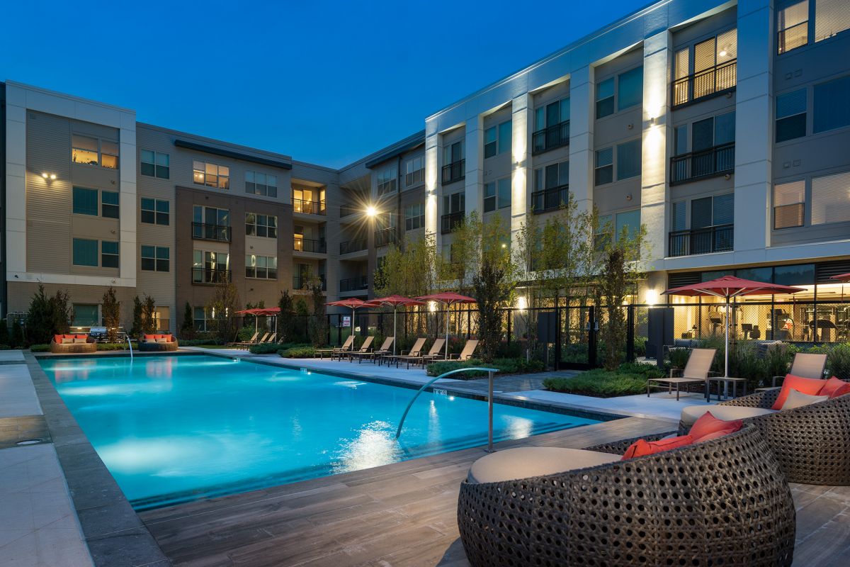 Modern apartment complex with a lit outdoor pool, lounge chairs, and woven seating at dusk.