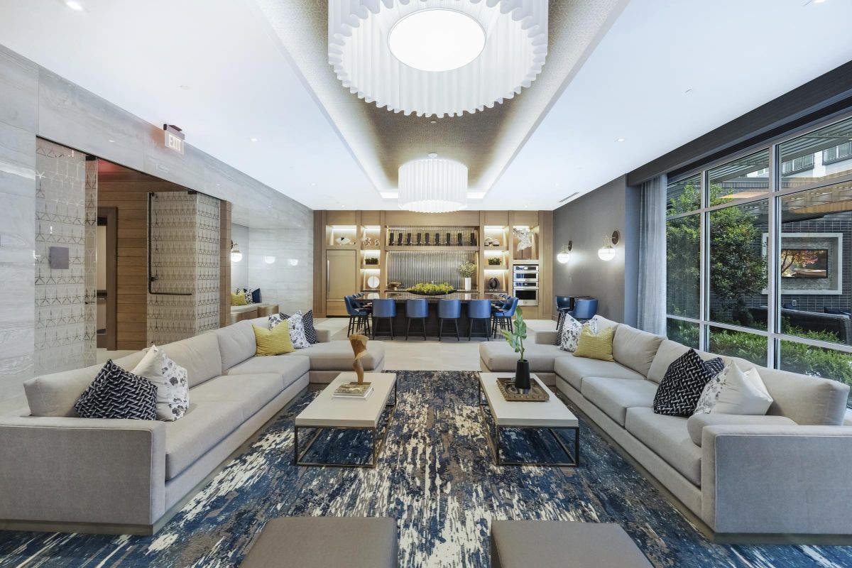 Modern living room with beige sofas, patterned cushions, a central rug, and large windows showing greenery.