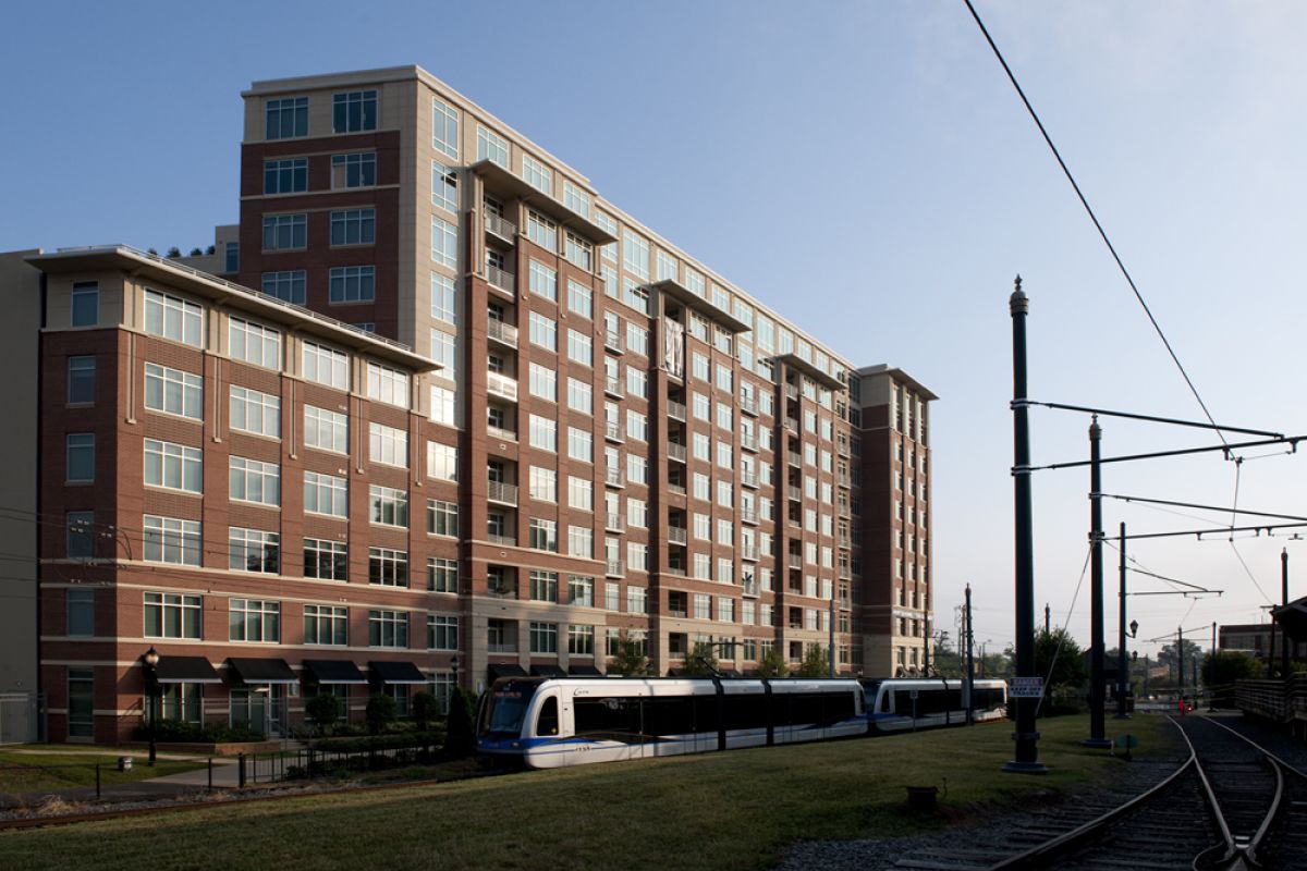 A light rail train passes a modern mid-rise apartment building on a sunny day.