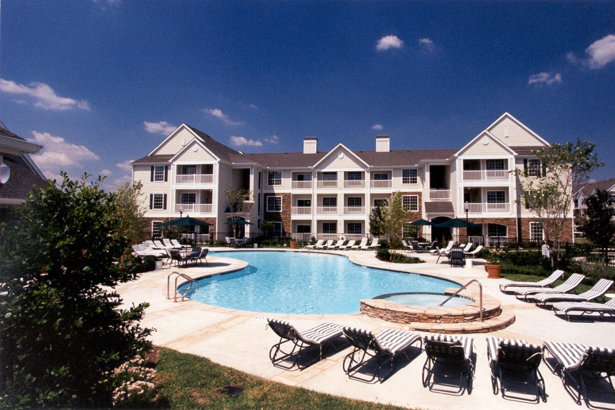 A modern apartment complex with a central swimming pool, lounge chairs, and landscaped greenery under a blue sky.