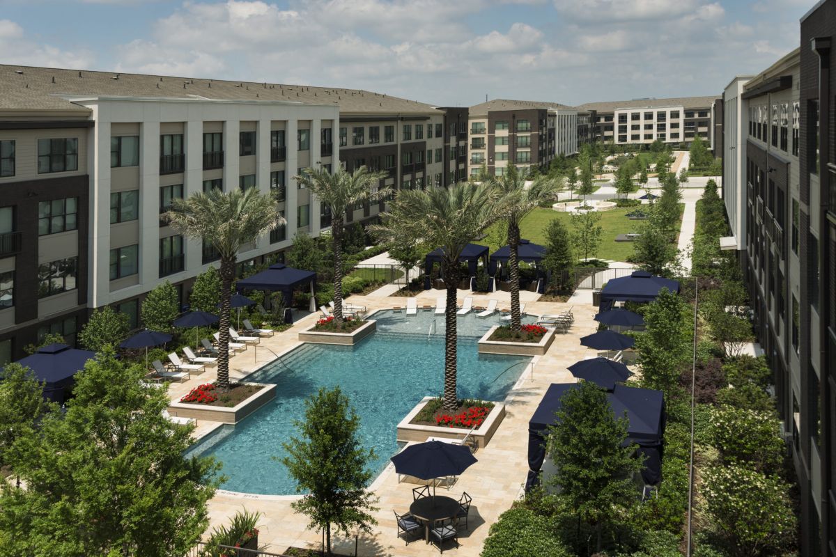 Large apartment complex with a central outdoor pool, palm trees, and lounge chairs, surrounded by green lawns.