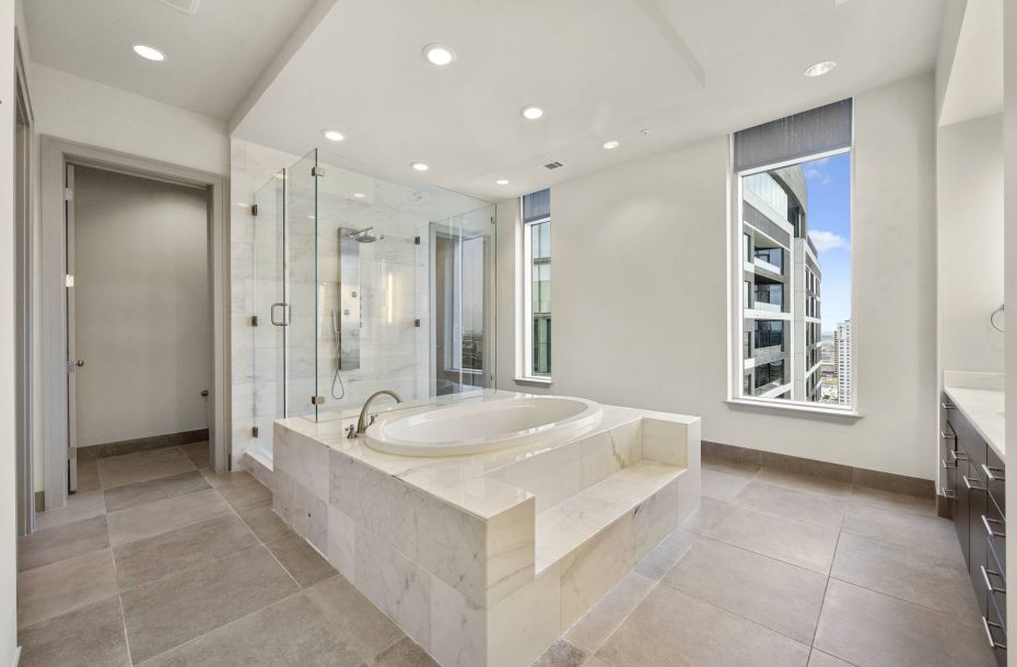 Hanover Company, Hanover Post Oak Modern bathroom with a central marble bathtub, glass shower, large windows, and gray tiled flooring.