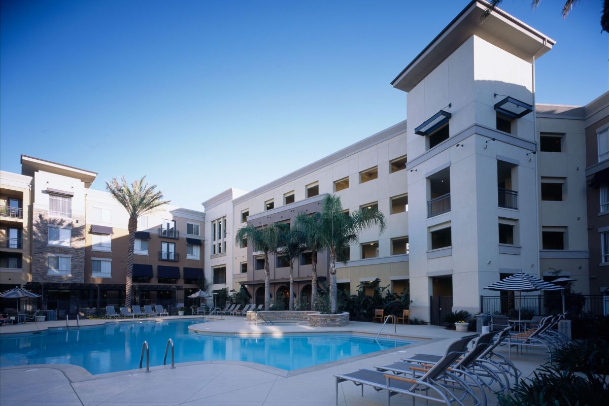 Outdoor swimming pool surrounded by lounge chairs and palm trees, next to a modern apartment building.