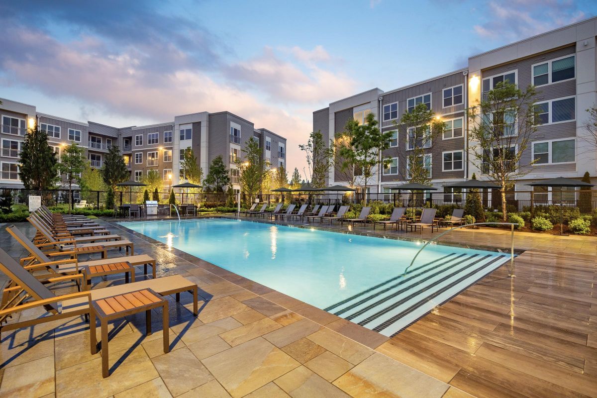 Modern apartment complex with a lit outdoor swimming pool, lounge chairs, and greenery at dusk.