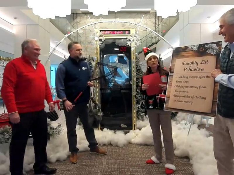 Four people stand indoors with holiday decor; one woman holds a sign listing "Naughty List Behaviors.
