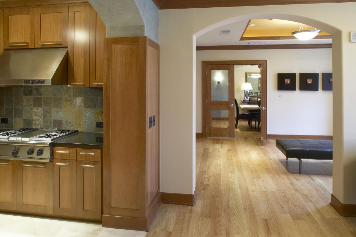 Modern kitchen with wooden cabinets opens to a hallway with light wood floors and a glimpse of a dining area.