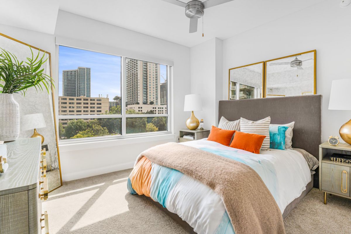 Modern bedroom with a large window, city view, decorative pillows, and lamps. Neutral tones with turquoise accents.