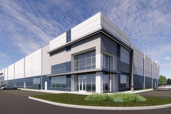 Modern industrial building with glass facade, surrounded by parking lot, cars, and landscaped greenery under a blue sky.