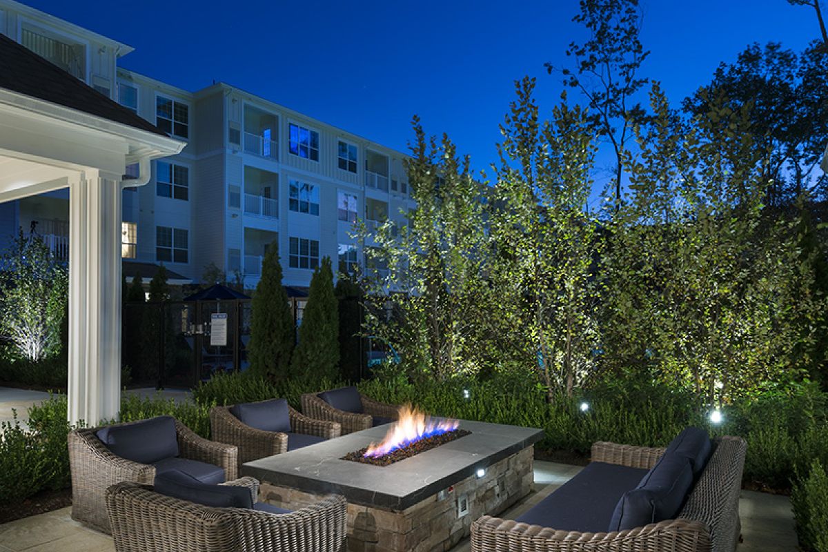 Wicker patio chairs surround a lit fire pit in a landscaped courtyard at dusk, with buildings in the background.
