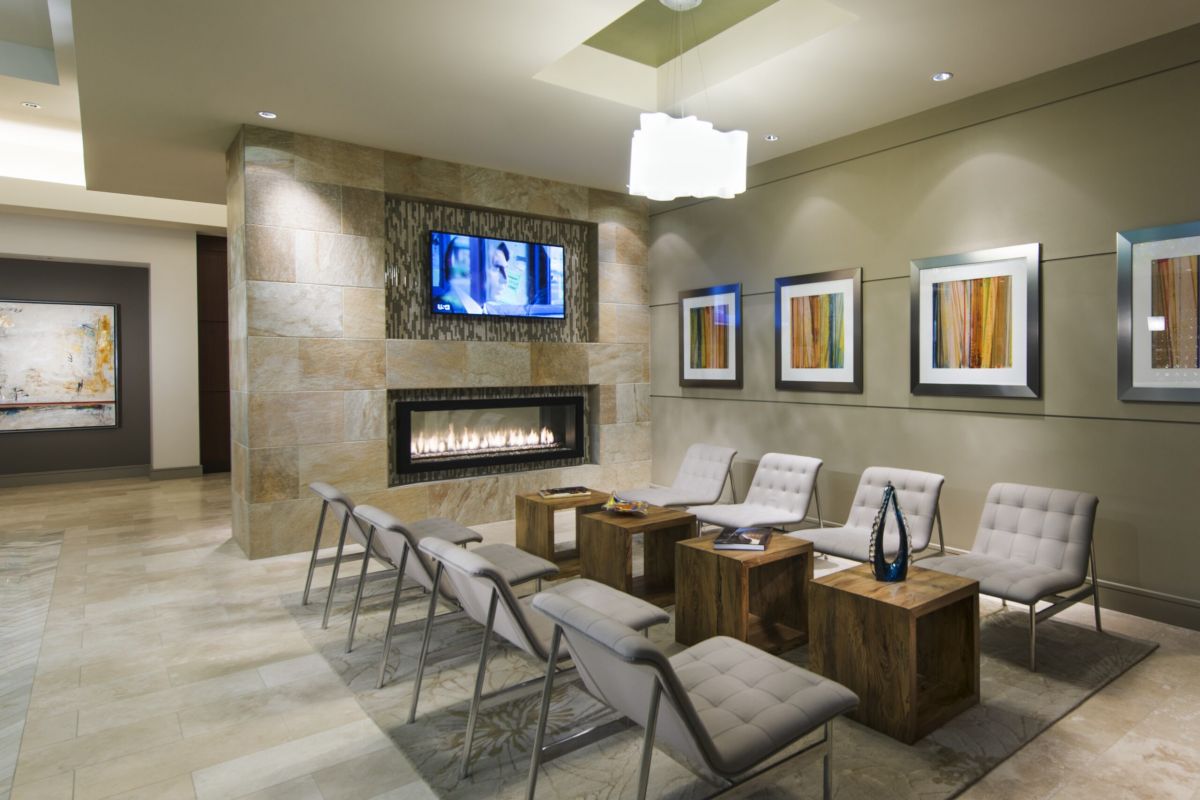 Modern waiting room with gray chairs, wooden tables, wall art, a fireplace, and a TV mounted above the fire.