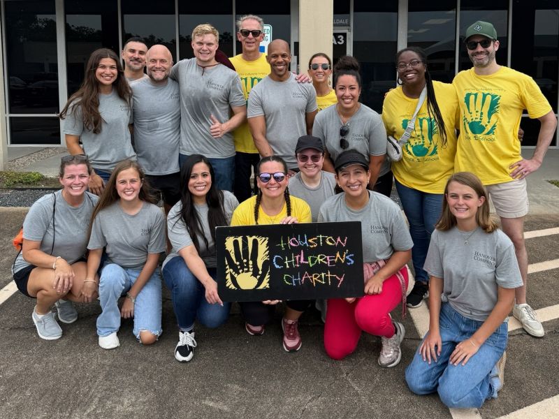 Group of smiling volunteers from Hanover Helping Hands pose outside with a "Houston Children’s Charity" sign, wearing matching shirts.