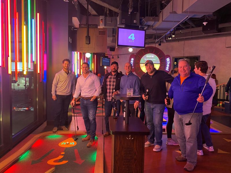 Seven men pose with mini golf clubs and drinks in a brightly lit indoor mini golf venue with neon lights.