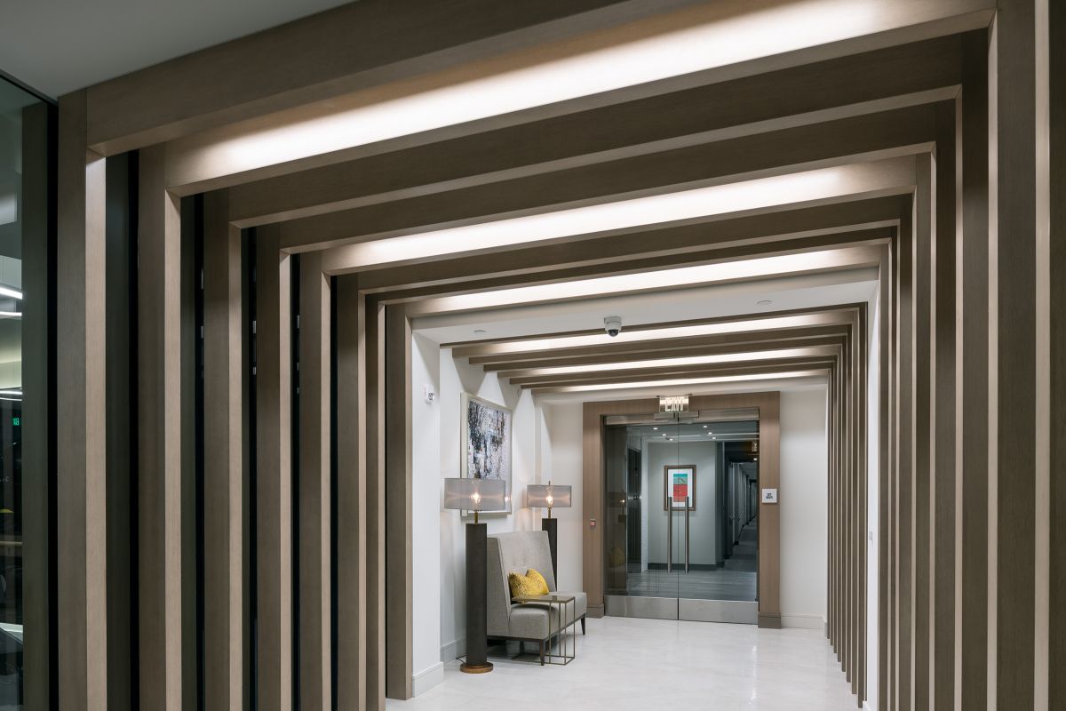 Modern hallway with wooden beams, seating area, artwork, lamps, and glass doors at the end.