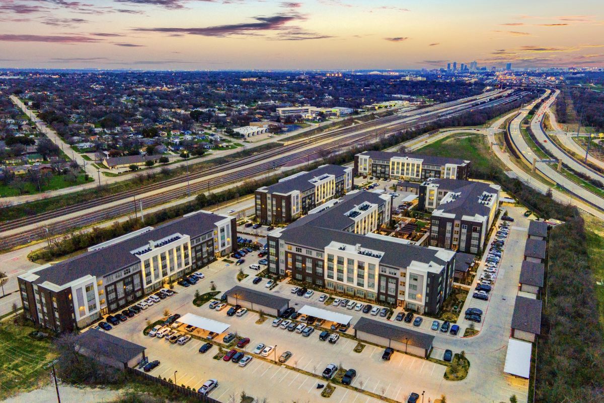 Aerial view of a residential complex with parking lots next to highways, with a city skyline in the distance at dusk.