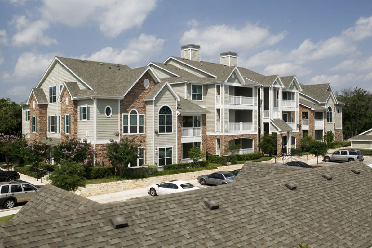 A modern, multi-story apartment building with balconies and parked cars in front, under a partly cloudy sky.