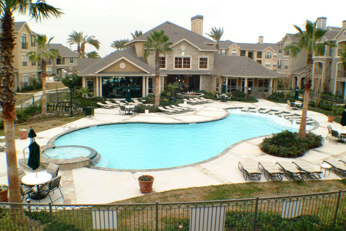 Large outdoor pool with lounge chairs, palm trees, and a clubhouse in a residential apartment complex.