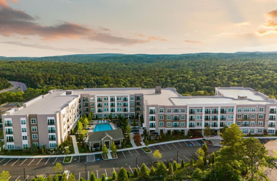 A modern apartment complex with a central pool, surrounded by trees and mountains under a cloudy sky.