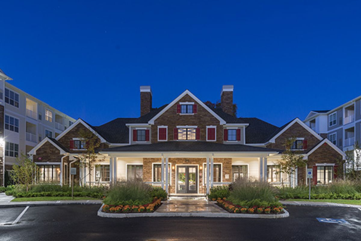 A well-lit apartment building with a central entrance and landscaped gardens at dusk.