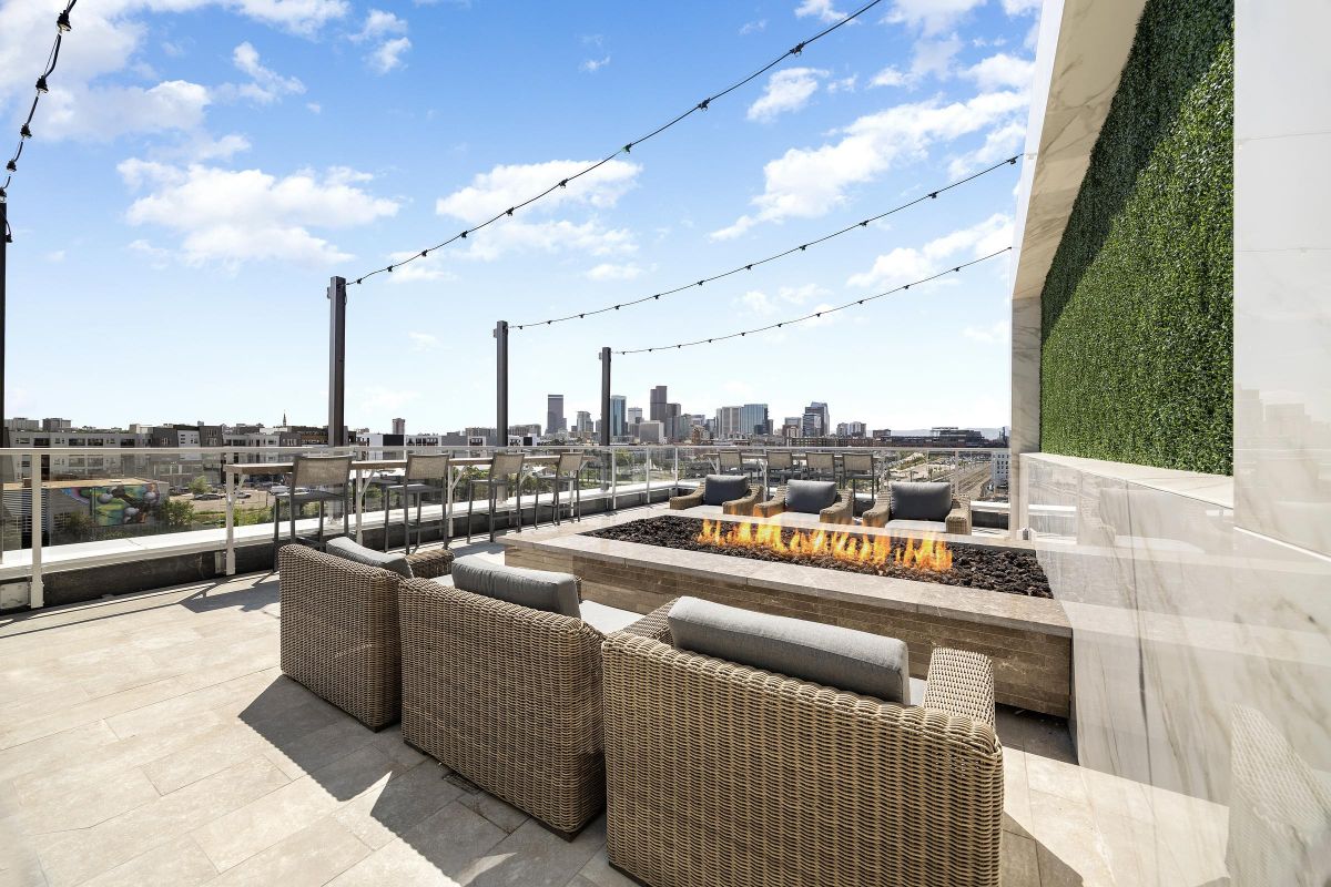 Rooftop patio with wicker chairs, a long firepit, city skyline view, and string lights under a blue sky.