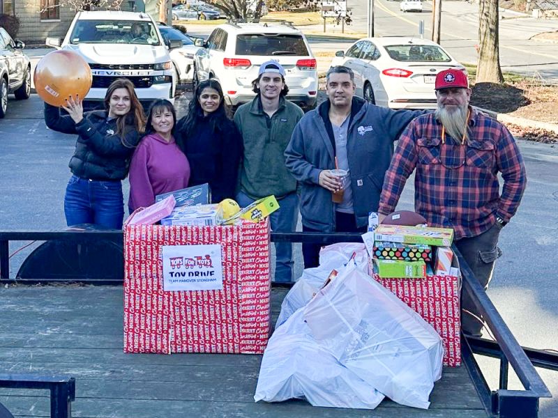 Six people stand behind wrapped boxes labeled "Toy Drive," filled with toys, on a trailer in a parking lot.