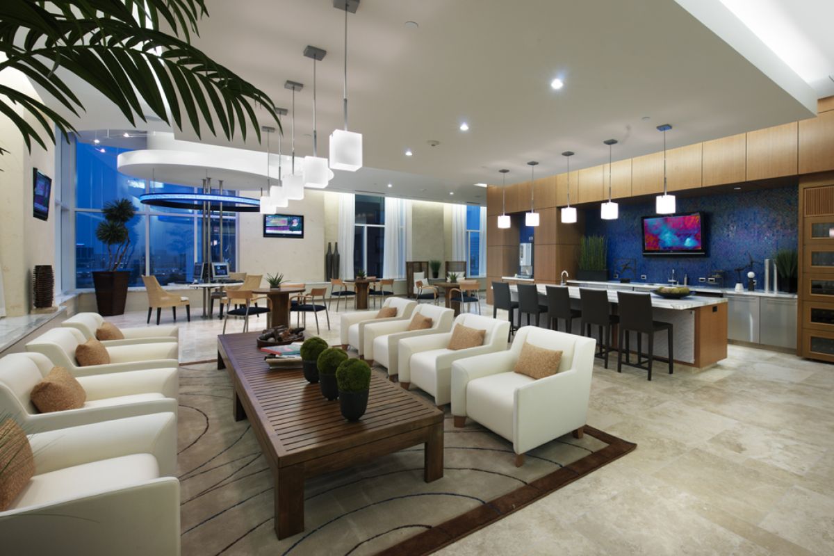 Modern, spacious lounge with white armchairs, wooden table, bar area, and large windows overlooking a city.