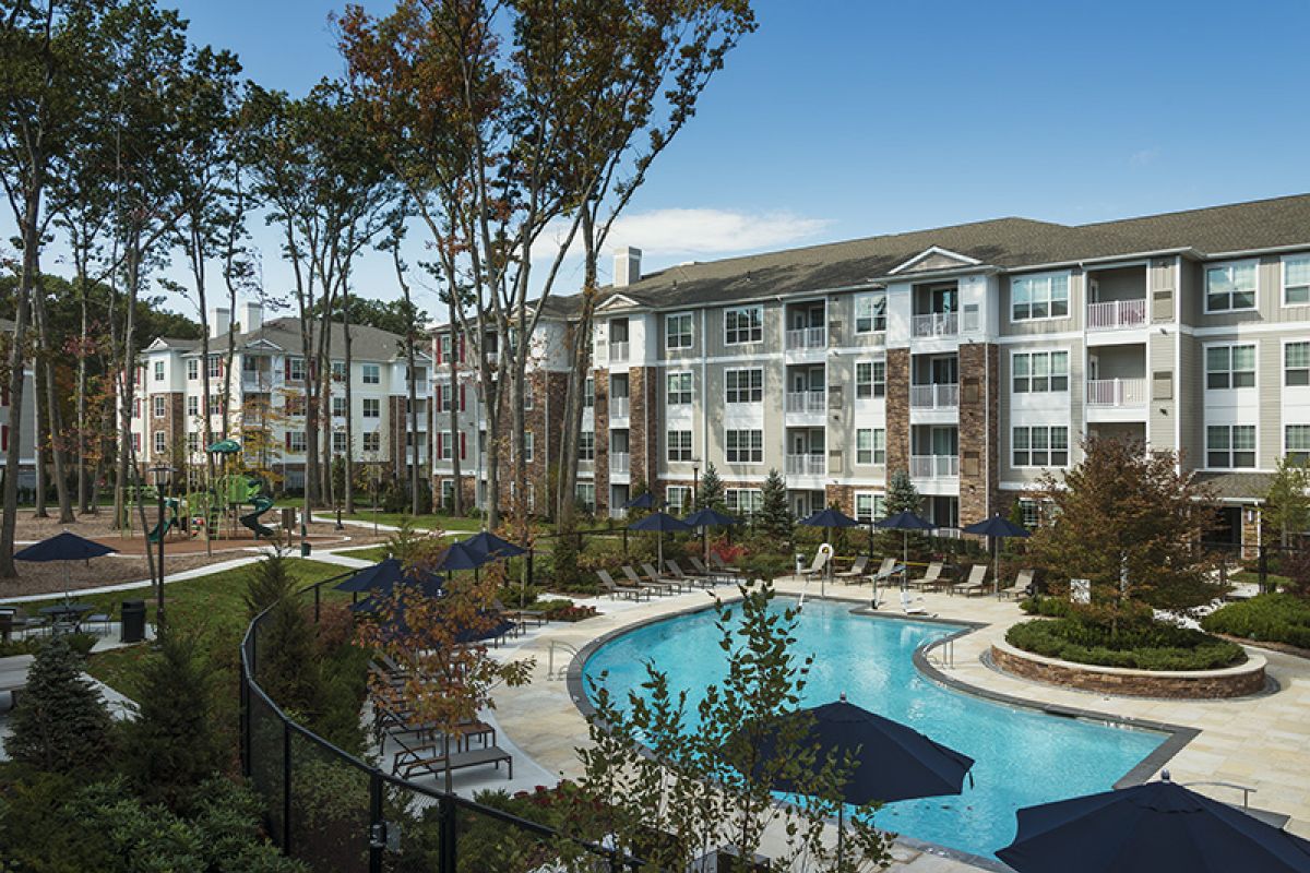 Modern apartment complex with a central outdoor swimming pool, surrounded by trees and lounge chairs.