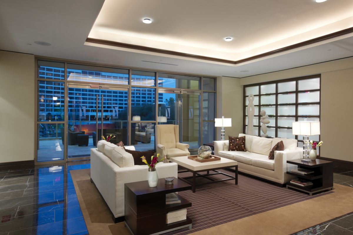 Modern living room with cream sofas, a brown rug, and large windows showing a view of buildings outside.