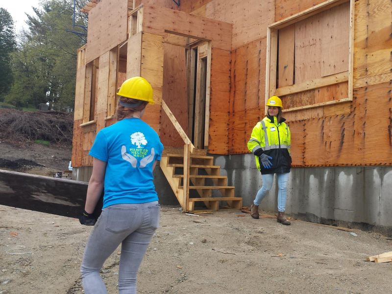 Two women in safety gear work near a wooden house under construction at a building site.