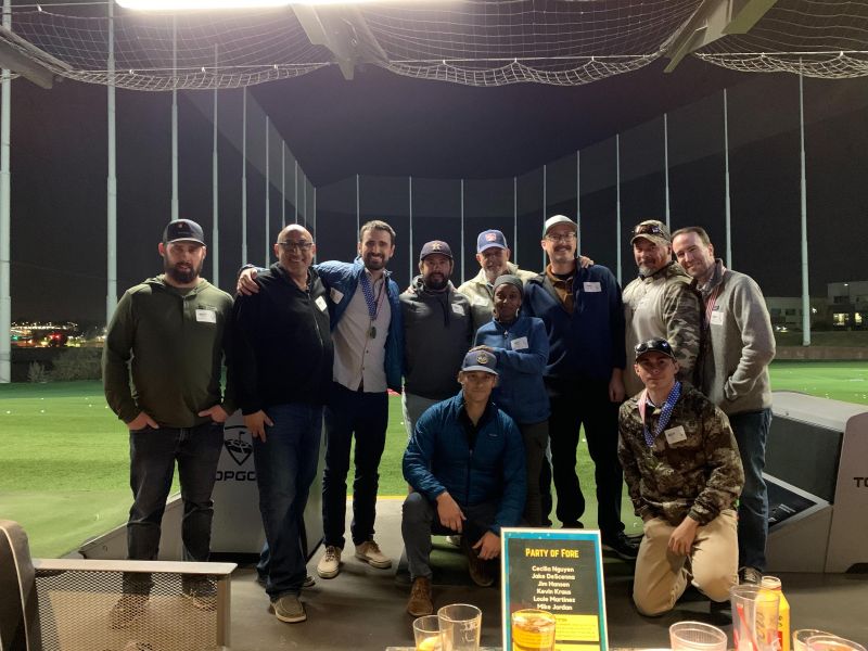 A group of eleven people pose together at a nighttime driving range with golf clubs and nets in the background.