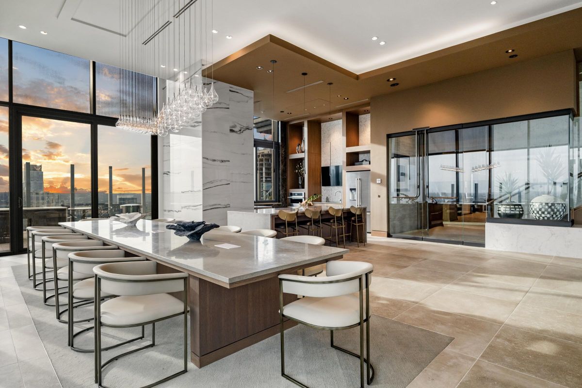 Luxurious modern kitchen and dining area with a large table, marble counters, and sunset view through tall windows.