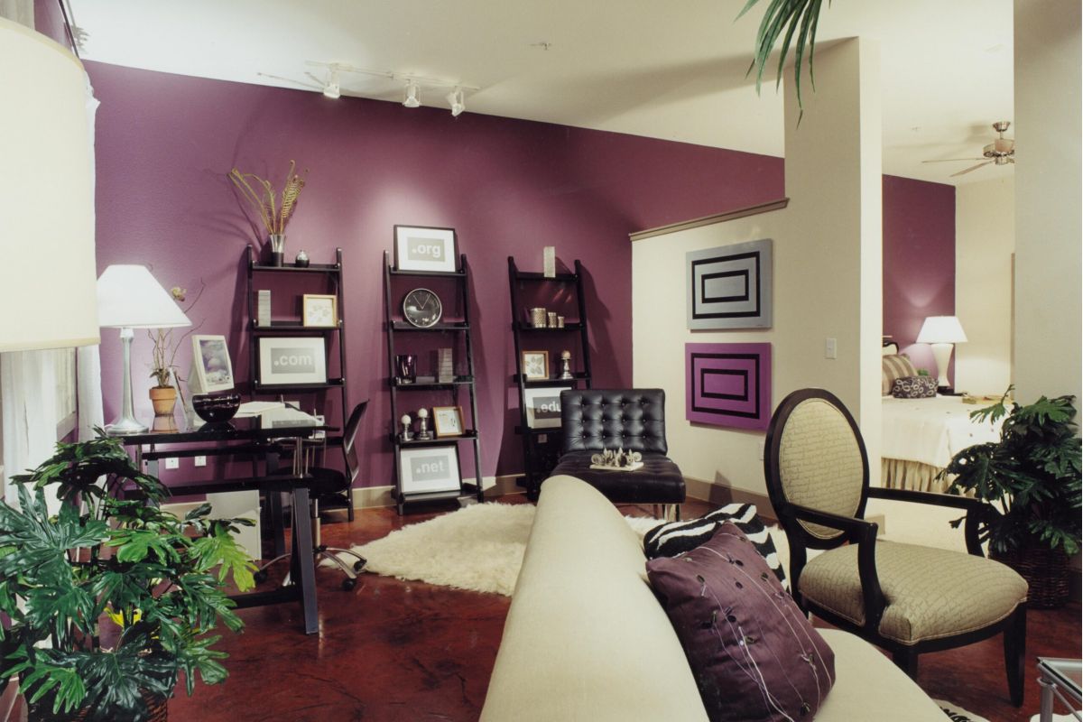 Modern living room with purple accent wall, shelves, desk, and beige furniture; plants decorate the space.