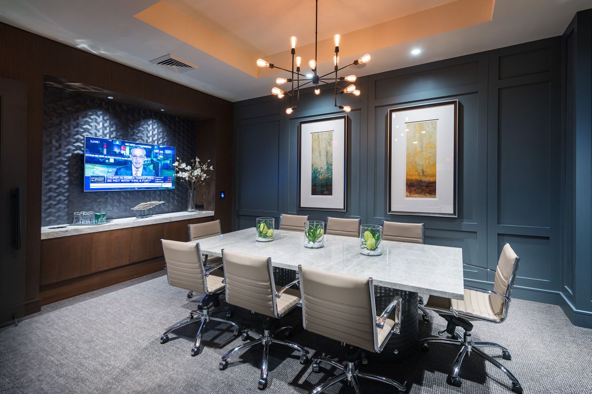Modern conference room with a marble table, beige chairs, wall art, and a TV displaying news.