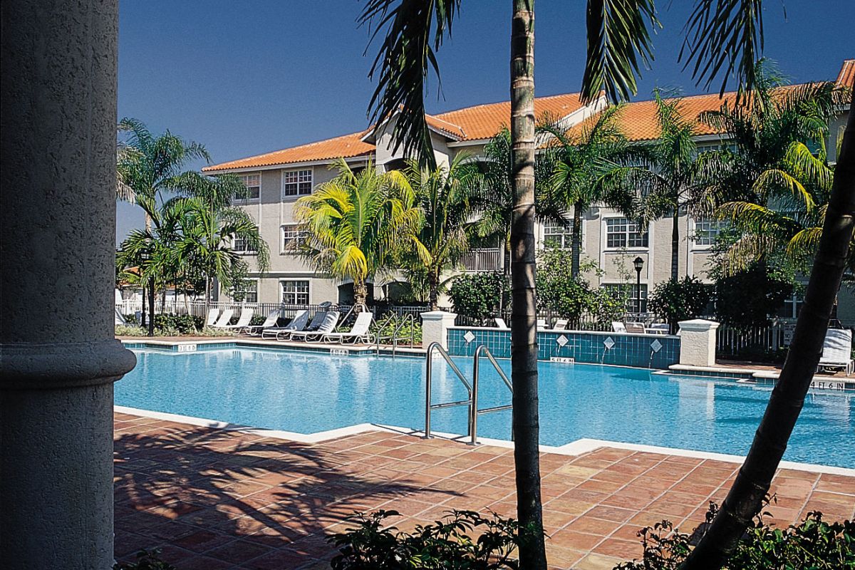 Sunny outdoor swimming pool at Broadwater Galveston, surrounded by palm trees and lounge chairs near a building with a red roof.