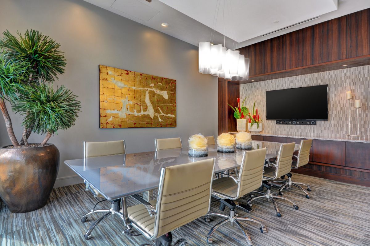 Modern conference room with a long table, beige chairs, plants, art, and a large wall-mounted TV.