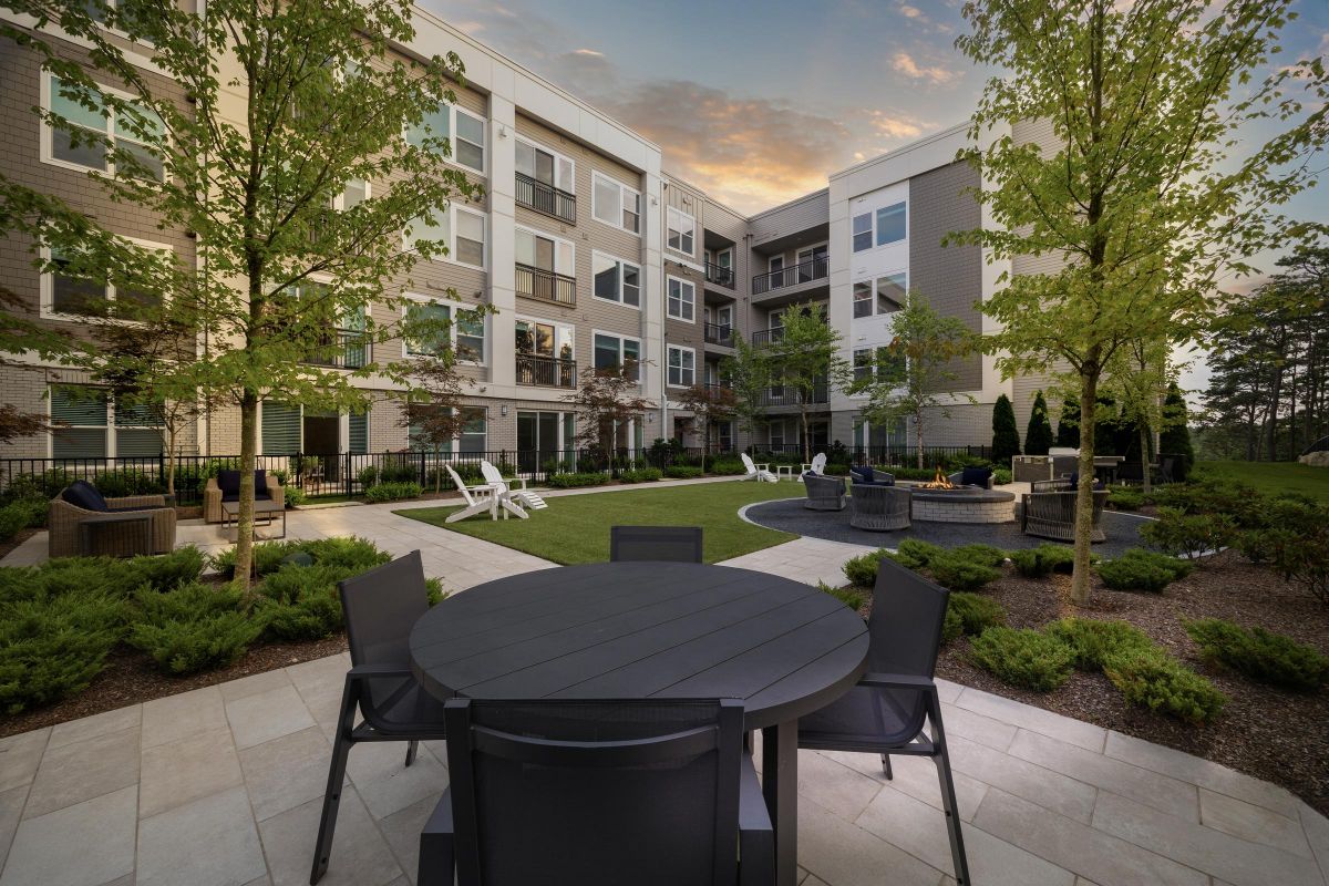 Modern apartment courtyard with trees, seating, fire pit, and surrounding apartments at sunset.