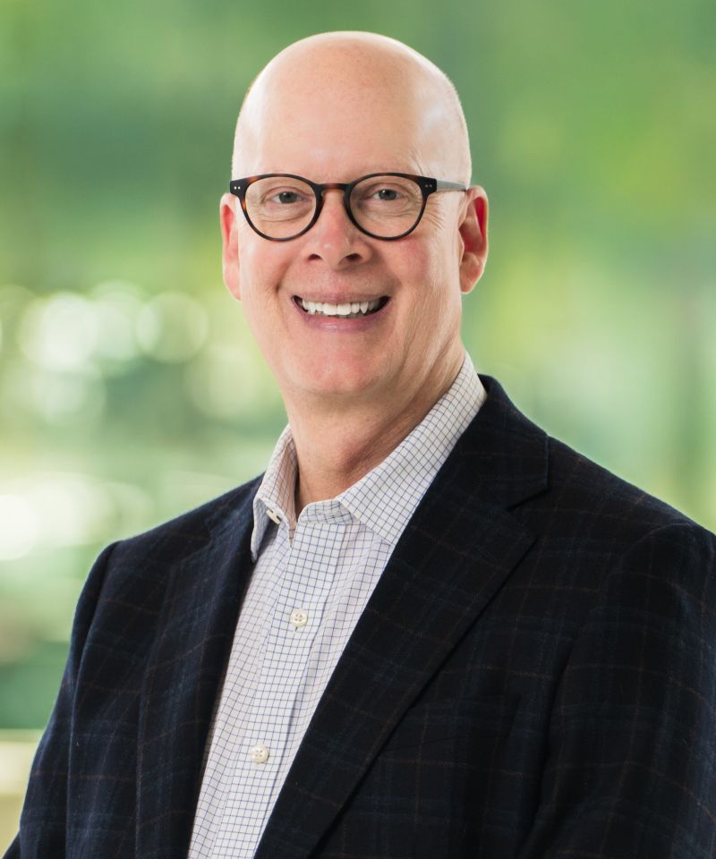 Smiling bald man with glasses in a suit jacket and checked shirt, standing in front of a blurred green background.