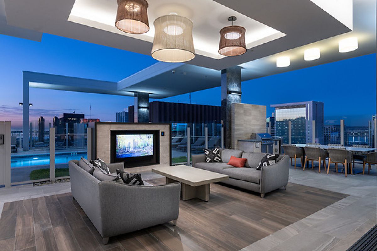 Modern rooftop lounge with gray sofas, a TV, dining area, and cityscape views at dusk.