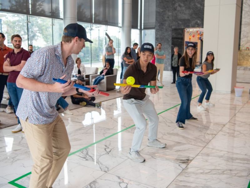 Three people race indoors balancing balls on paddles, with others watching and cheering in the background.