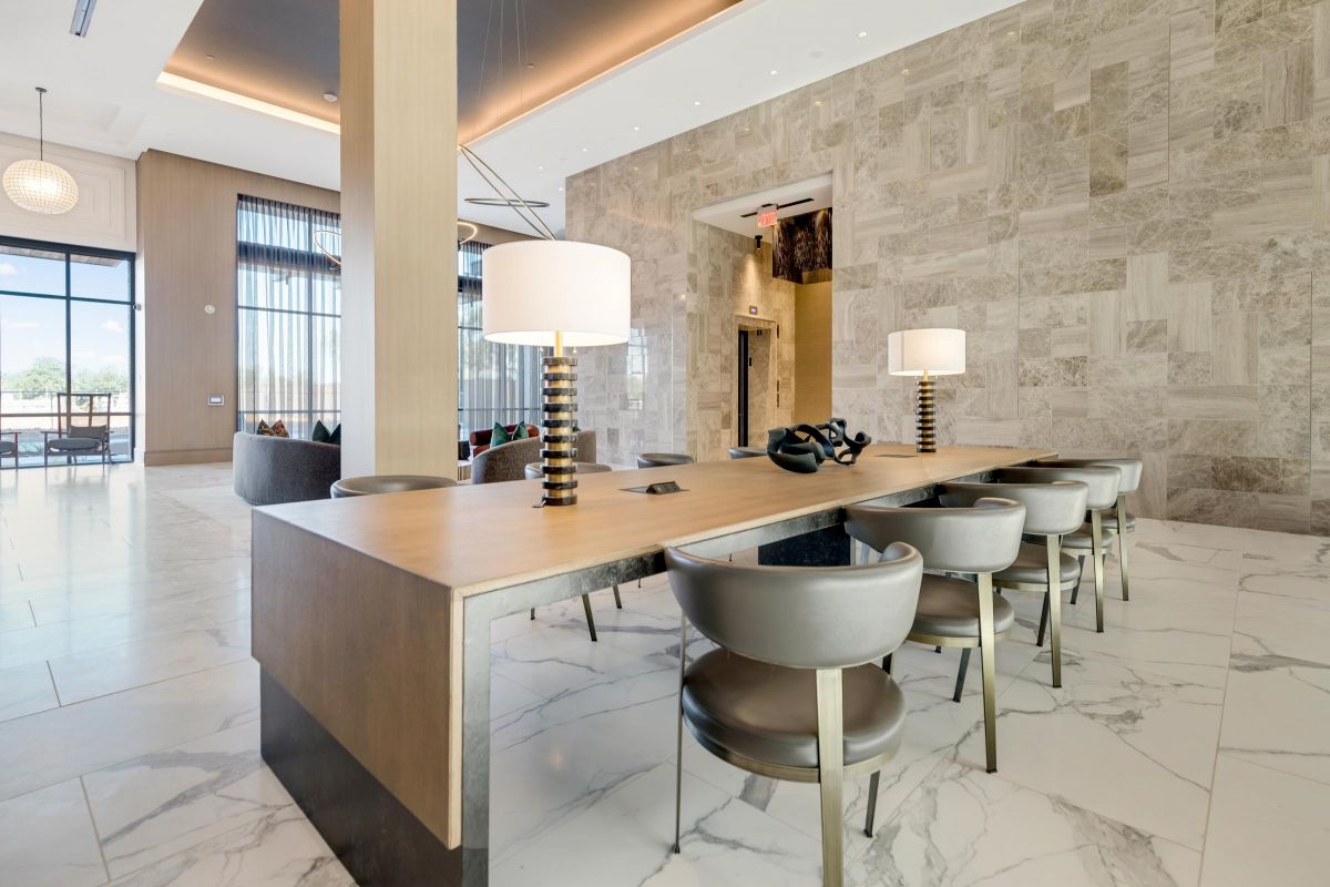 Modern lounge area with a long wooden table, gray chairs, and two lamps on marble tile floor.